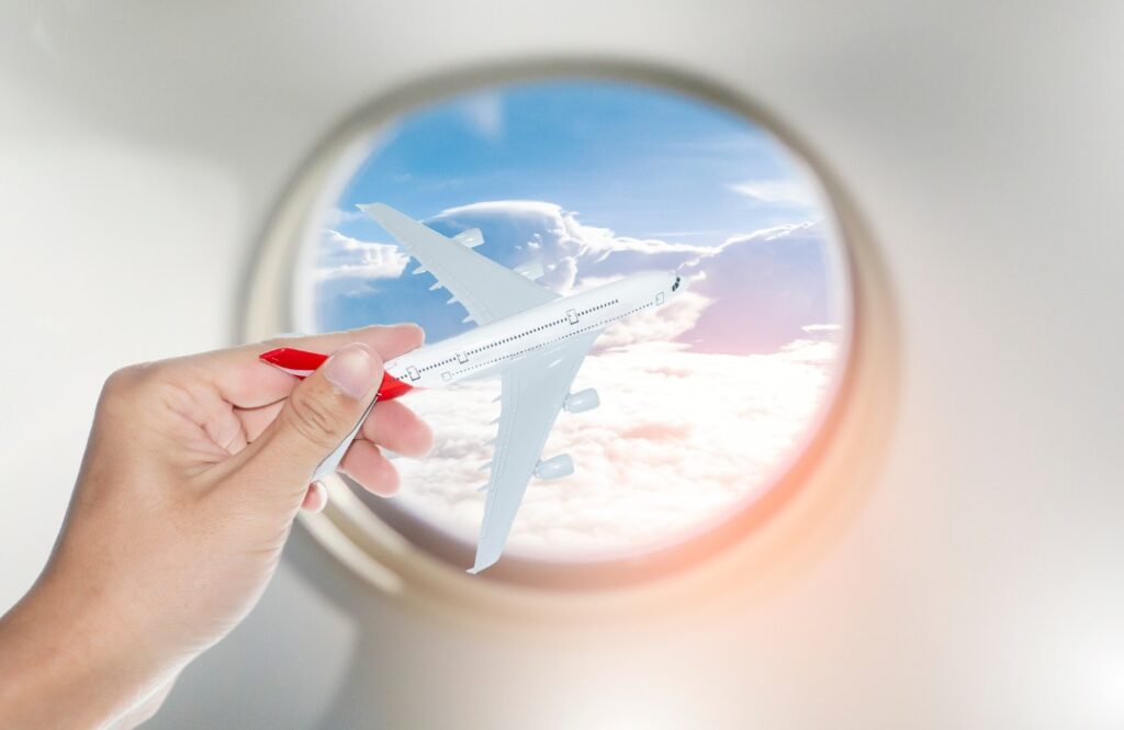 A close-up of a hand holding a model airplane next to a plane window, with clouds and the sky in the background, representing travel planning and tips on how to find cheap flights for budget-friendly journeys.
