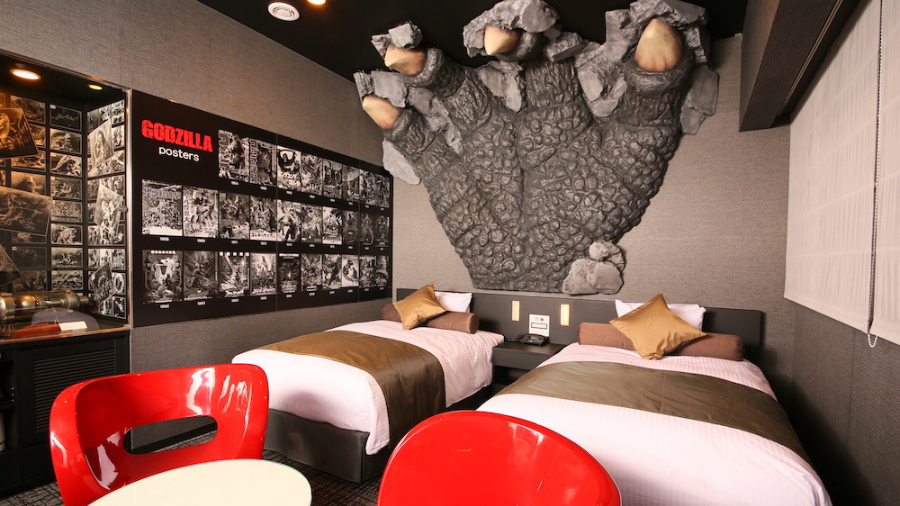 A themed hotel room with twin beds, a large Godzilla claw coming out of the wall, and a collection of Godzilla posters.