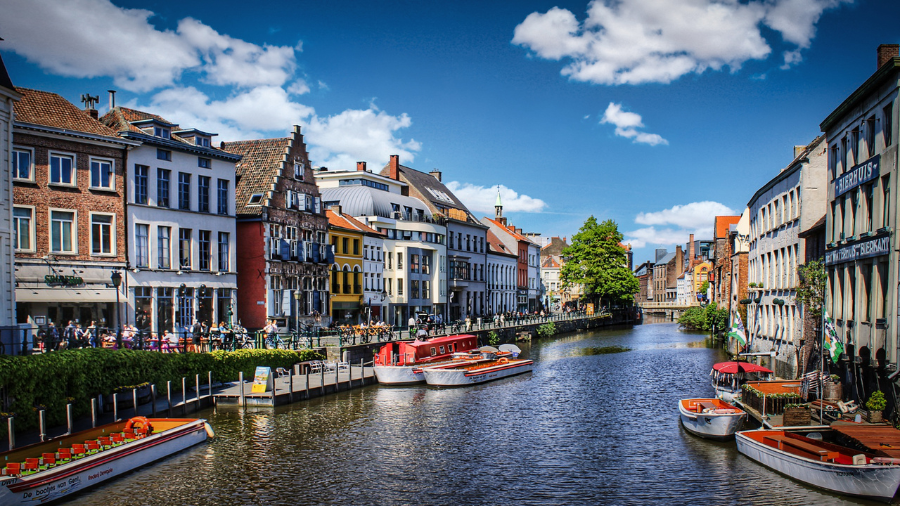 A scenic canal view in a European city with colorful historic buildings, boats docked along the water, and a bright blue sky with clouds.