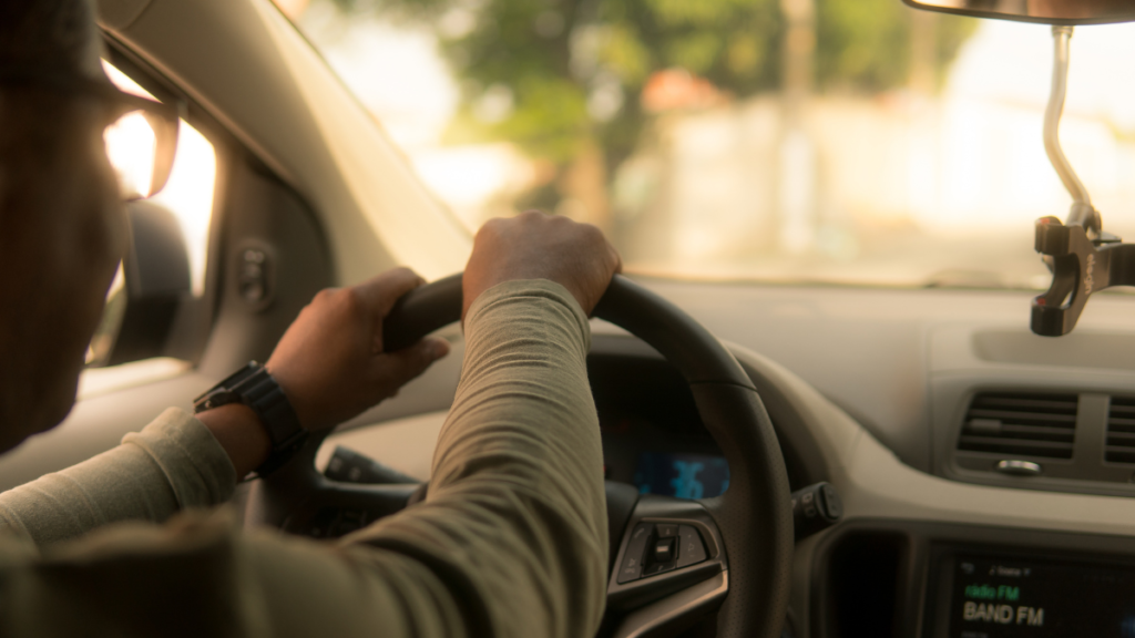 A close-up of a man's hands on the steering wheel, driving through a sunlit road.