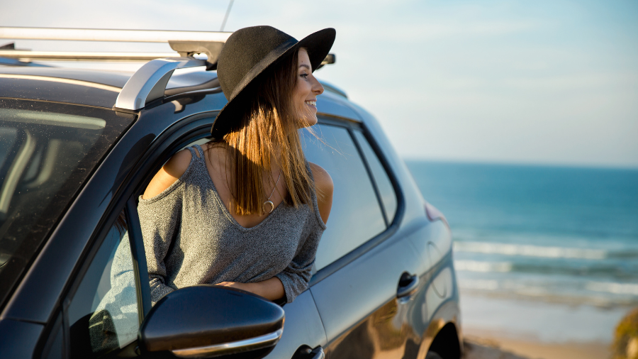 A young woman wearing a black hat and gray sweater leans out of a car window, smiling as she enjoys the ocean view.