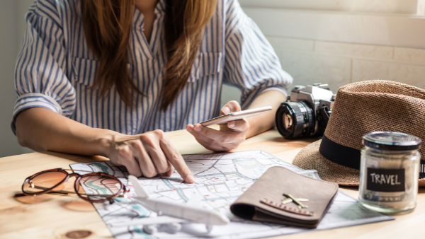 A woman planning a trip using a smartphone and a map, with travel essentials like a camera, passport, and hat on the table.